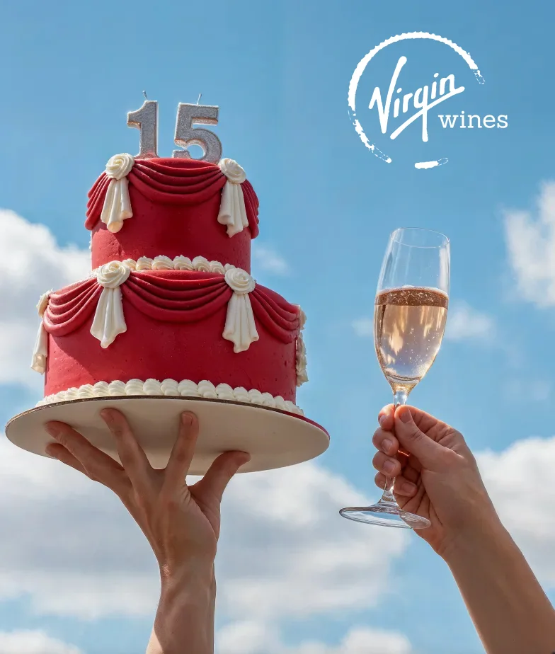 A red fifteenth birthday cake held up against a blue sky beside a raised glass of sparkling wine, with the Virgin Wines logo
